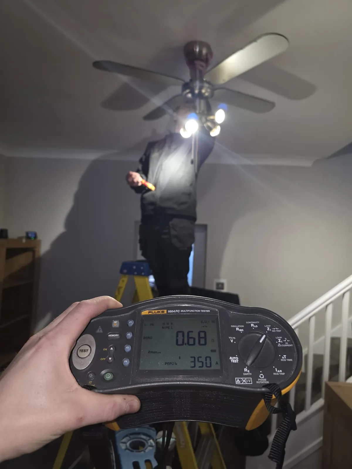 Testing a ceiling fan light from a ladder with the multifunction tester reading in the foreground